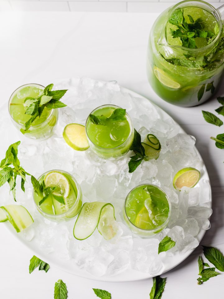 a pitcher filled to the top with cucumber ginger mint agua fresca along with four small glasses filled to the top and garnished with sliced limes, mint leaves, and cucumber slices. The glasses are on an oval platter filled with ice while the pitcher is on a flat surface in the upper right hand corner